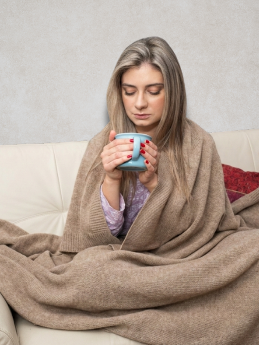 A woman wearing the Rayaa cashmere blanket in brown color sitting on a sofa with a coffee mug

